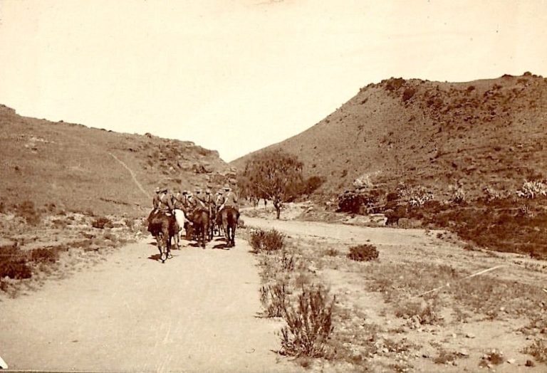 A sepia-toned landscape featuring a dirt road and a group of people on Horseback walking away.