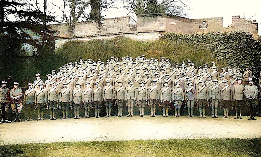 A large group of uniformed soldiers standing outdoors, with trees and a building in the background.