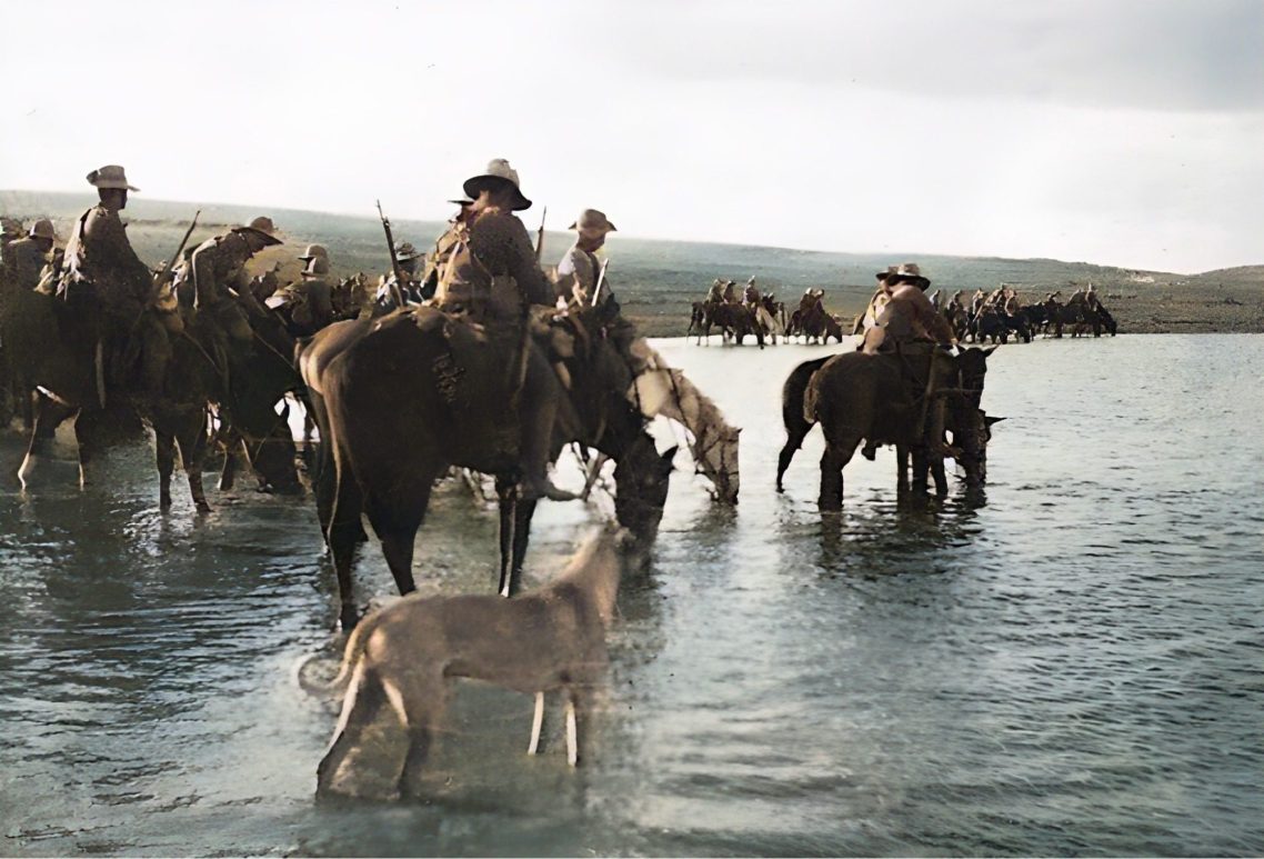 Cavalry on horseback wade through water, accompanied by a dog.
