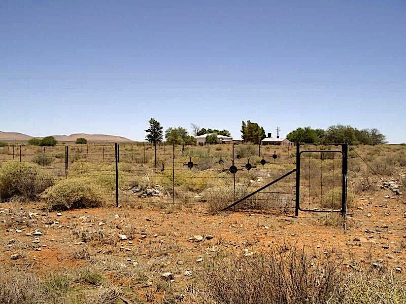 A rusty gate opens onto a dry landscape with sparse vegetation and distant hills.