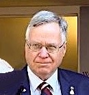 A man with glasses and grey hair, wearing a dark suit and tie, looks directly at the viewer.