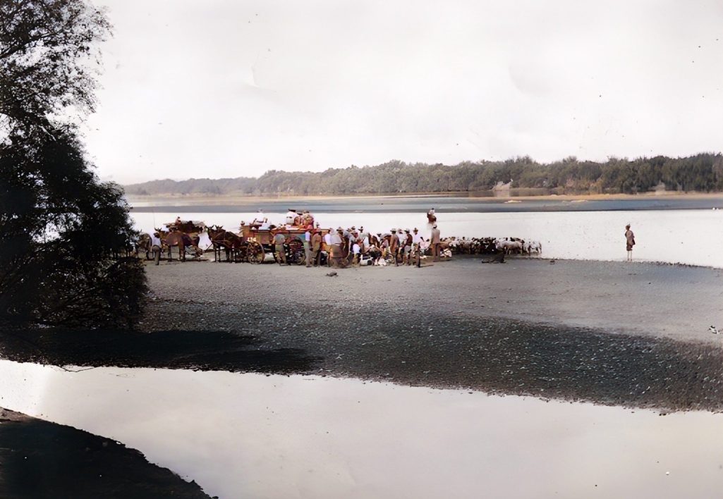 A sandy riverbank with people gathered, trees in the background, and calm water.