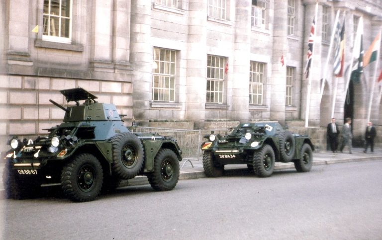Two military vehicles parked outside a historic building, with flags and people in the background.