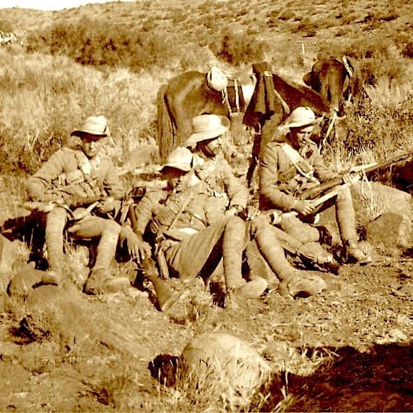 Four soldiers seated on rocks in a sepia-toned landscape, armed with rifles.