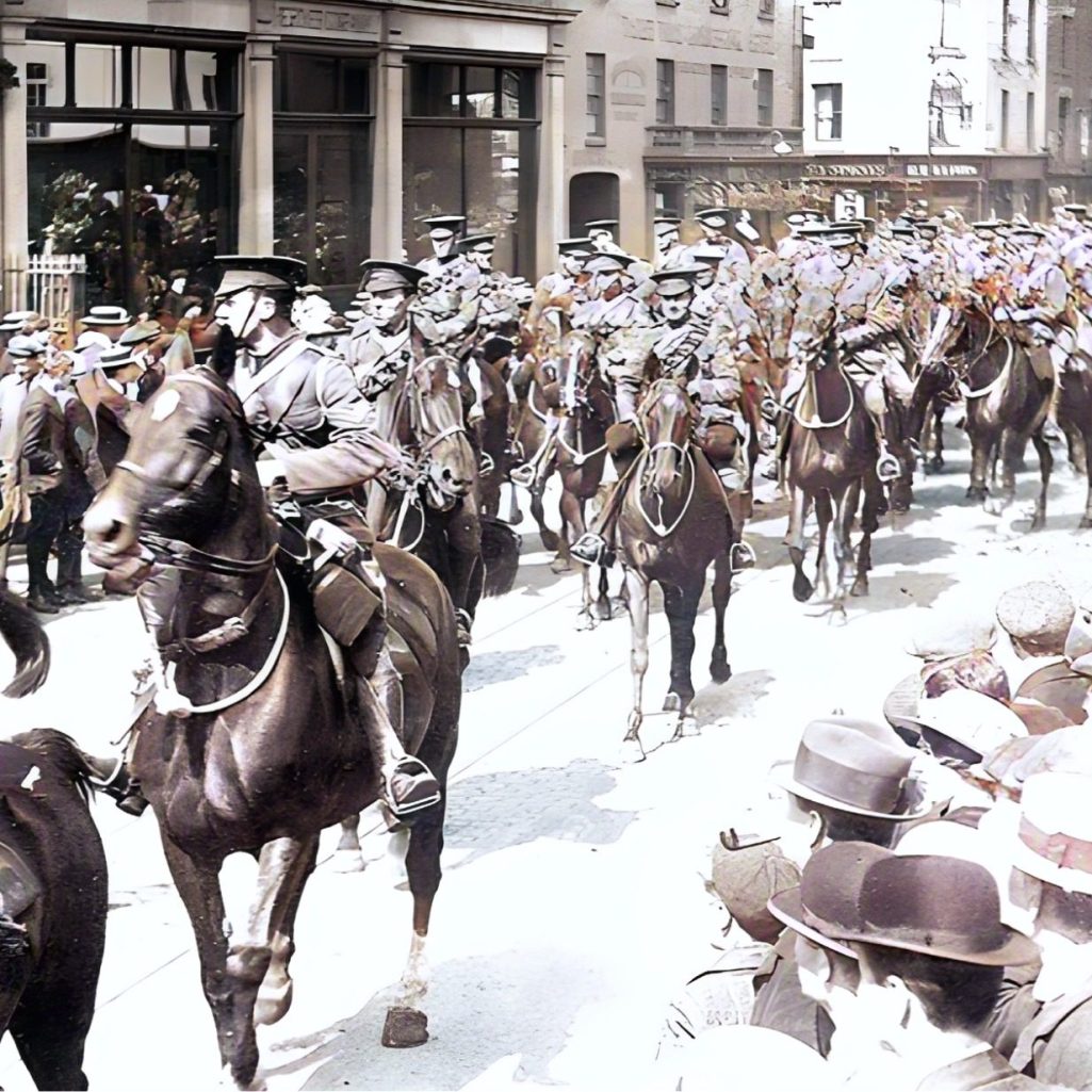 Crowd watching a mounted parade on a city street, with uniformed soldiers on horseback.