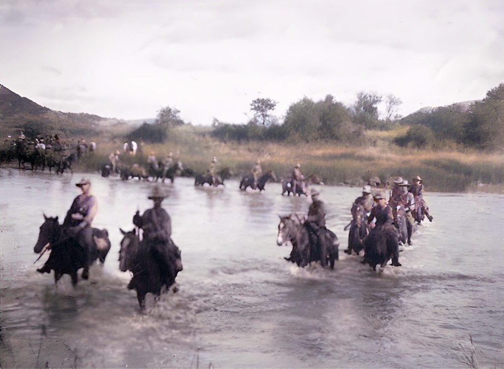 Troopers on horseback crossing a shallow river, surrounded by greenery and hills.